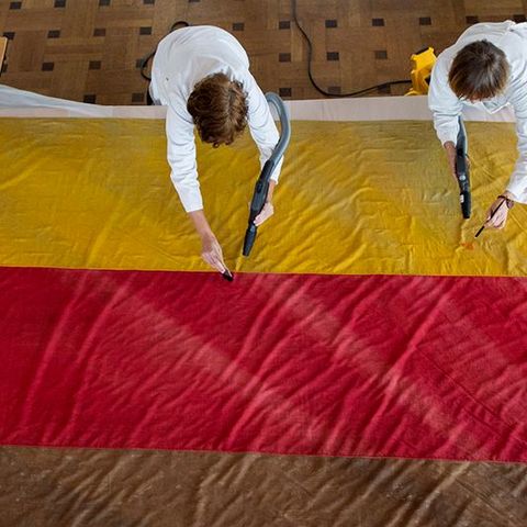 Auf dem Bild sieht man zwei Personen in weißen Arbeitskitteln, die eine große Stofffahne auf dem Boden reinigen bzw. restaurieren. Die Fahne ist Gelb, Rot und Braun gefärbt. Die Personen arbeiten mit kleinen Geräten.