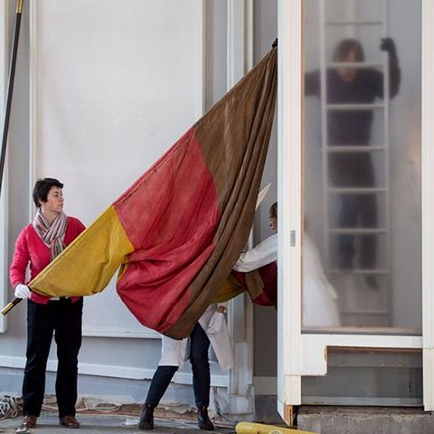 Auf dem Bild tragen mehrere Personen eine große rot-gelb-braune Fahne durch den Plenarsaal. Eine Person hält eine lange Stange, während andere beim Transport helfen. Im Hintergrund sieht man eine Leiter.