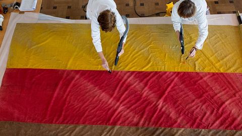 Auf dem Bild sieht man zwei Personen in weißen Arbeitskitteln, die eine große Stofffahne auf dem Boden reinigen bzw. restaurieren. Die Fahne ist Gelb, Rot und Braun gefärbt. Die Personen arbeiten mit kleinen Geräten.