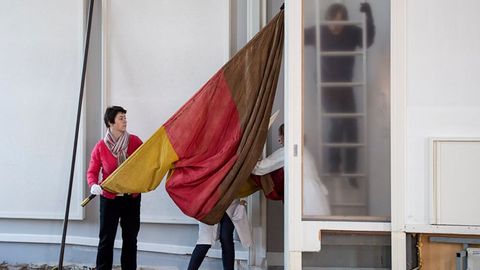 Auf dem Bild tragen mehrere Personen eine große rot-gelb-braune Fahne durch den Plenarsaal. Eine Person hält eine lange Stange, während andere beim Transport helfen. Im Hintergrund sieht man eine Leiter.