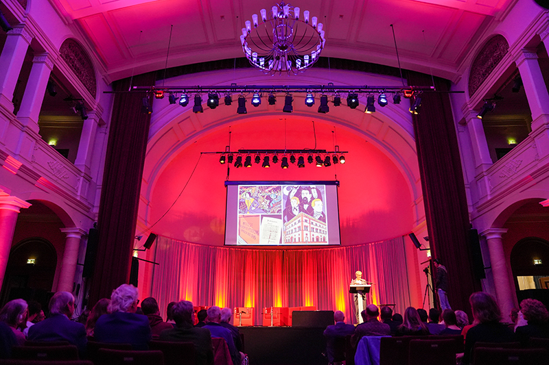 Landtagspr&auml;sident Hendrik Hering steht an einem Rednerpult auf einer B&uuml;hne in der Fruchthalle Kaiserslautern. Vor der B&uuml;hne sitzt ein Publikum. 