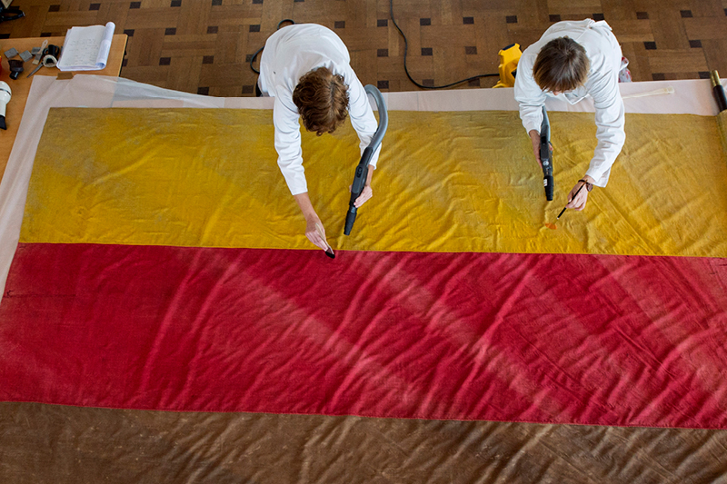 Auf dem Bild sieht man zwei Personen in weißen Arbeitskitteln, die eine große Stofffahne auf dem Boden reinigen bzw. restaurieren. Die Fahne ist Gelb, Rot und Braun gefärbt. Die Personen arbeiten mit kleinen Geräten.