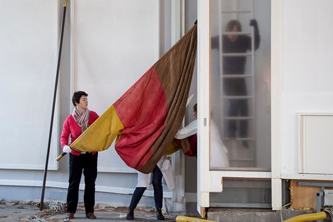 Auf dem Bild tragen mehrere Personen eine große rot-gelb-braune Fahne durch den Plenarsaal. Eine Person hält eine lange Stange, während andere beim Transport helfen. Im Hintergrund sieht man eine Leiter.