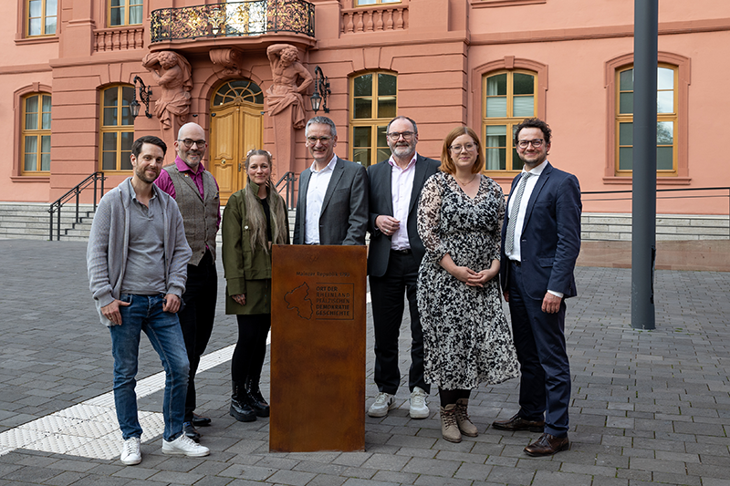 Hendrik Hering und die G&auml;ste des Abends stehen vor der k&uuml;rzlich verliehenen Auszeichung im Hof des Deutschhauses. Auf der bronzenen S&auml;ule steht: "Ort der rheinland-pf&auml;lzischen Demokratiegeschichte"